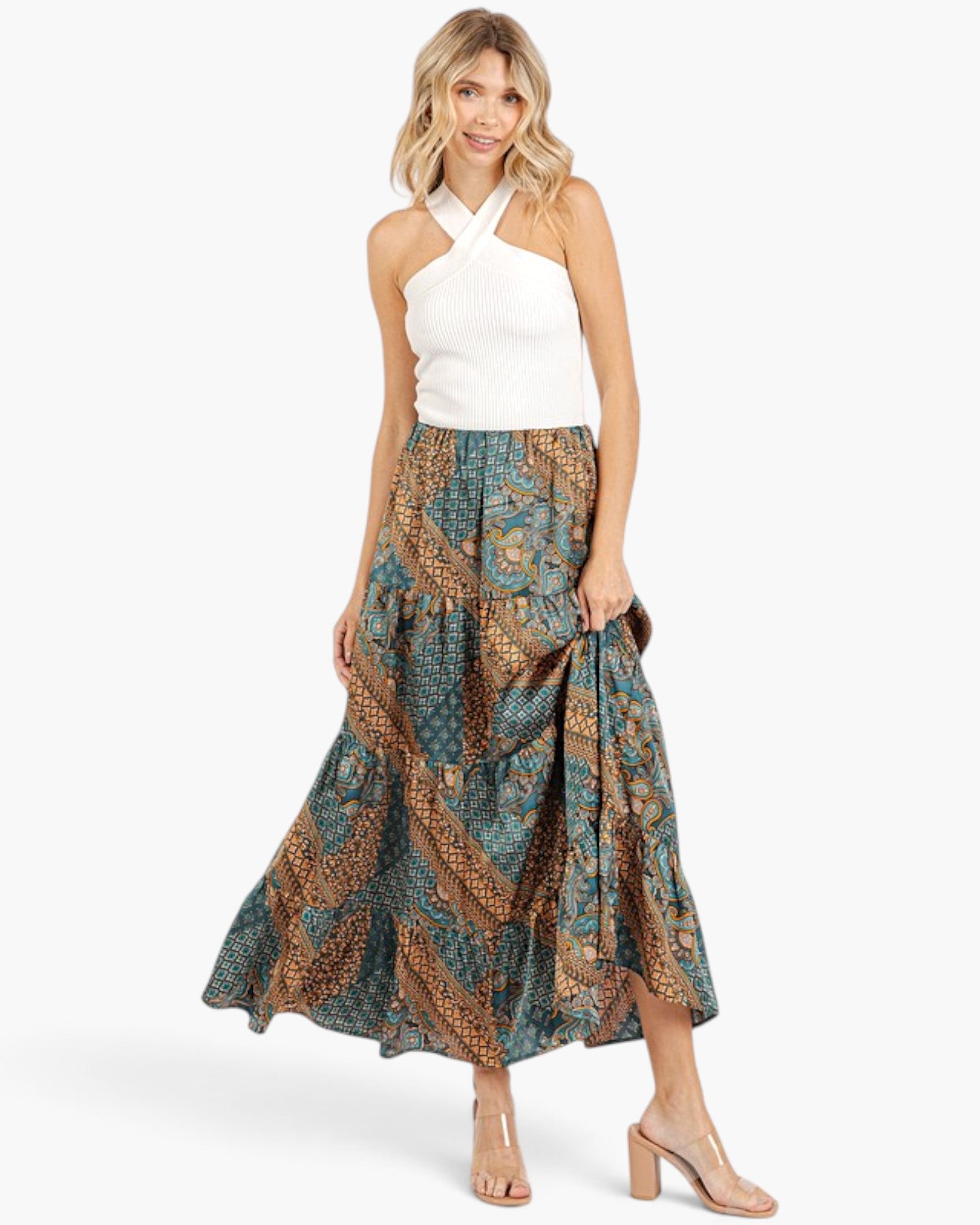 A woman modeling a white halter top and a long tiered maxi skirt with a brown and teal batik-style pattern, paired with clear block-heel sandals, against a light gray studio background.