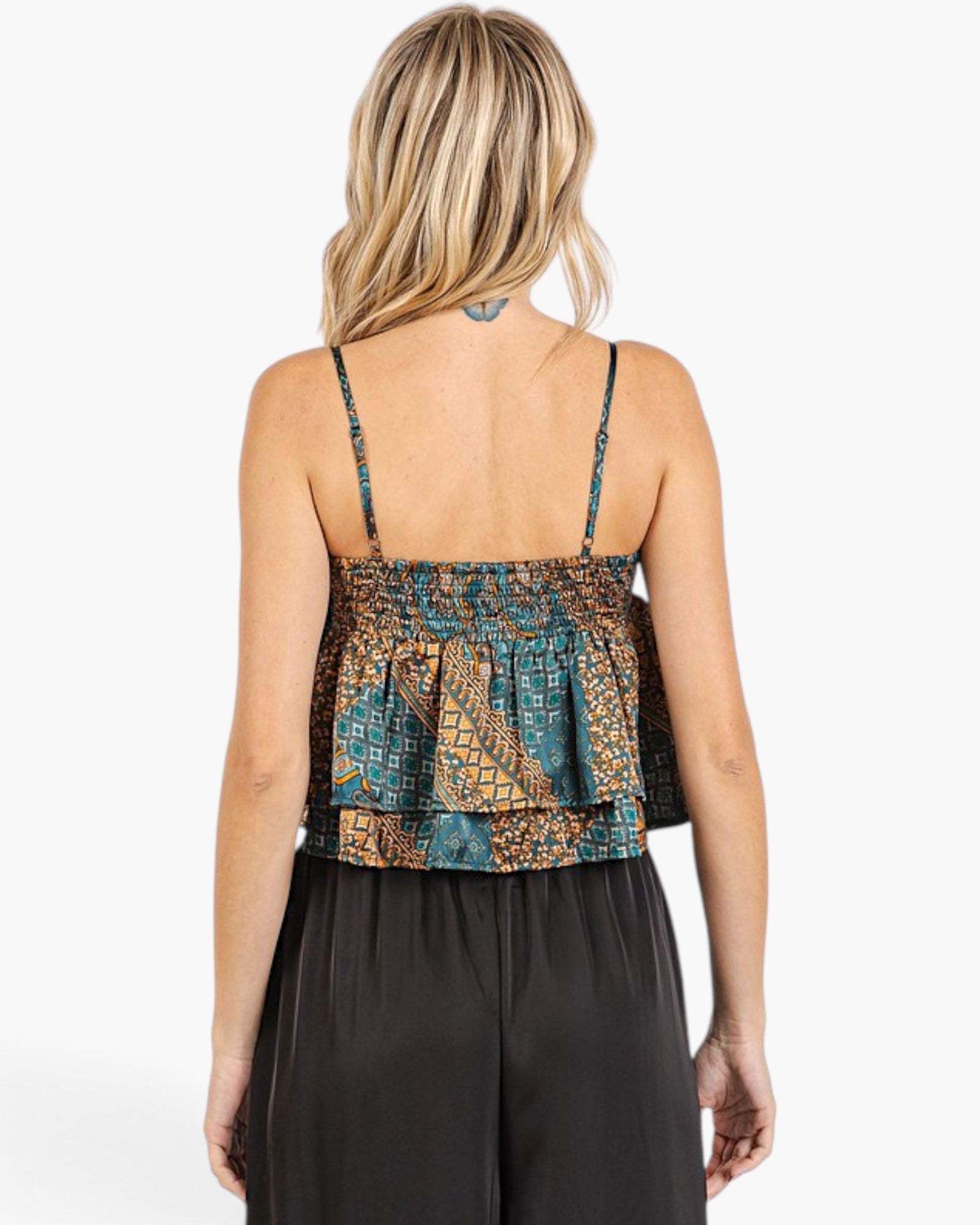 A woman shown from the back wearing a patterned camisole top with thin straps and a ruffled layered hem, paired with black wide-leg pants, against a plain white studio background.