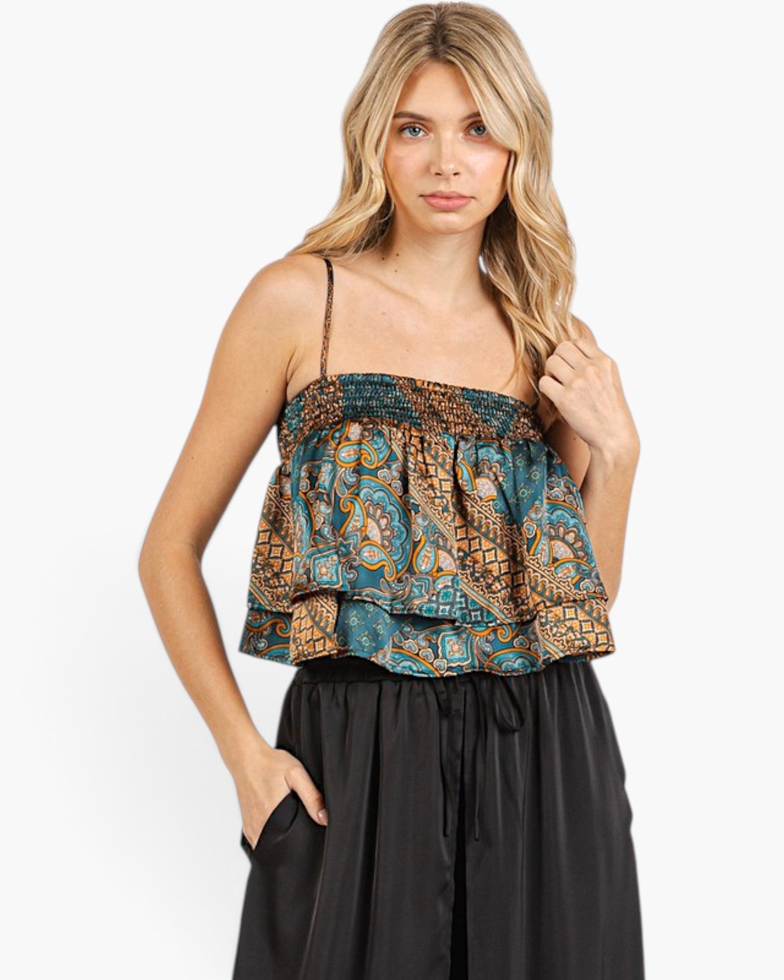 A woman modeling a sleeveless ruffled top with a blue, gold, and brown paisley pattern, paired with black wide-leg pants, against a plain white background.