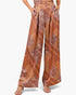 A model wearing high-waisted, wide-leg trousers with a brown, orange, and beige paisley pattern, paired with beige heeled sandals against a light gray background.
