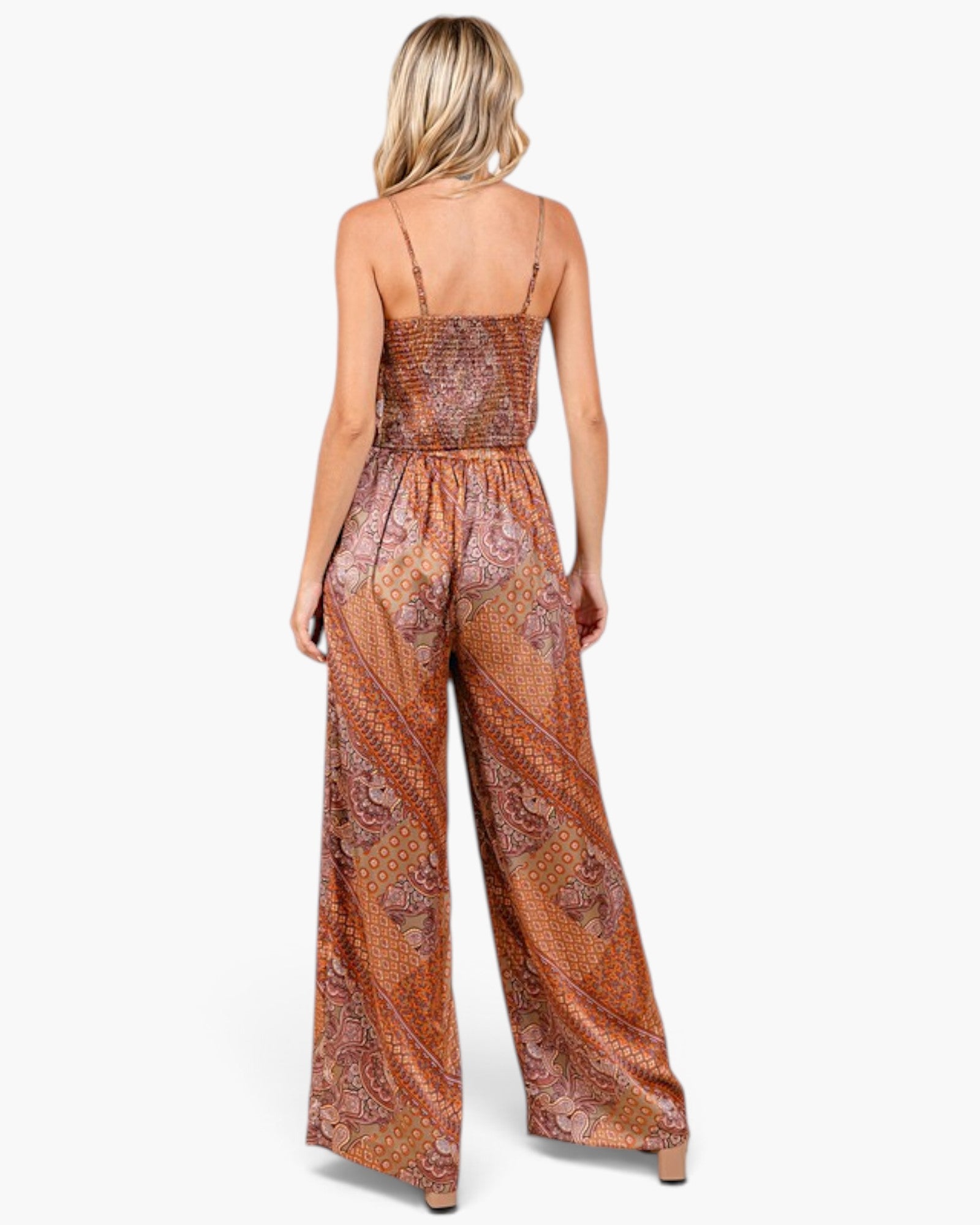 A woman viewed from the back wearing a sleeveless wide-leg jumpsuit with a smocked bodice and an orange-brown paisley pattern, set against a plain white background.