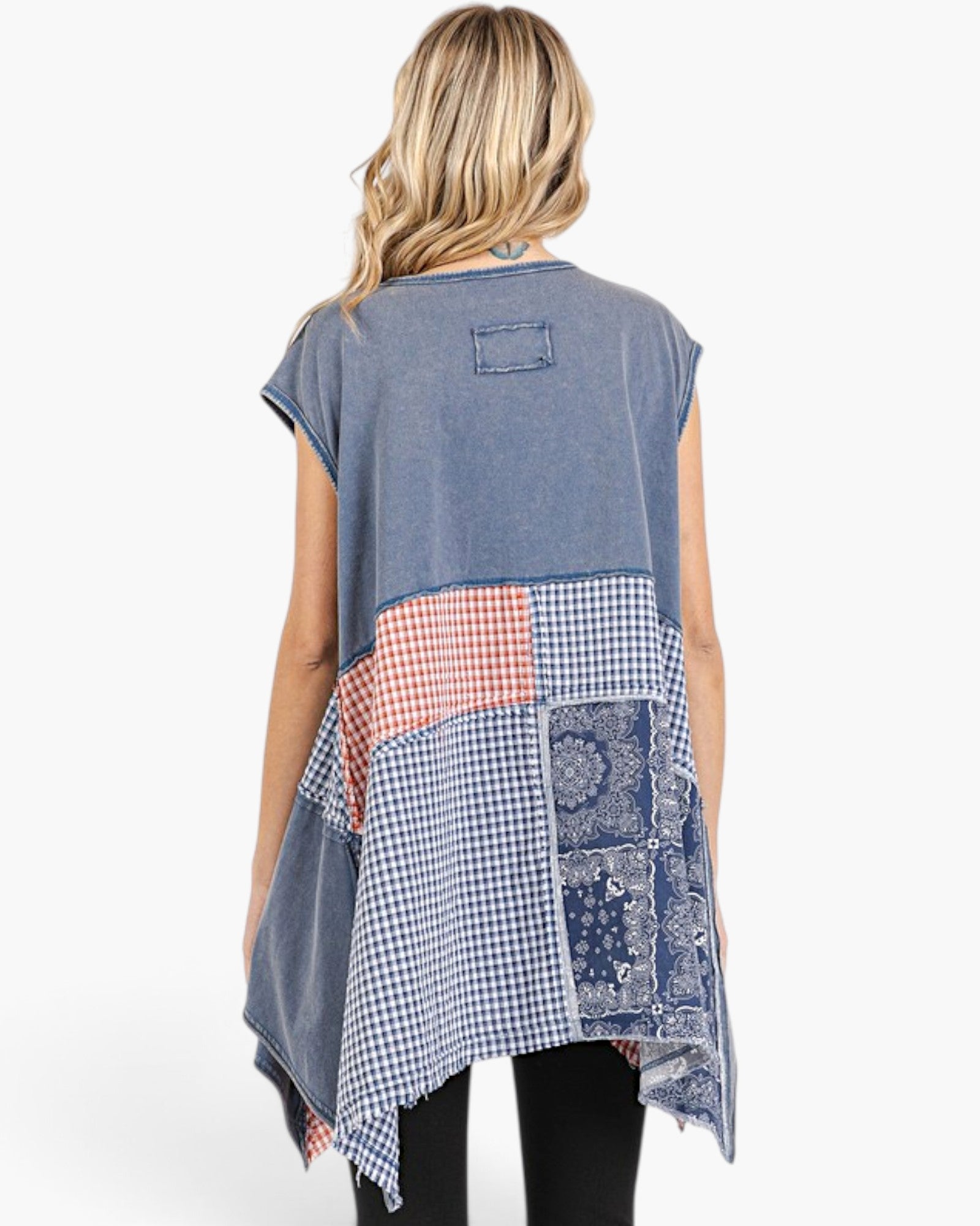 A woman viewed from the back wearing a sleeveless patchwork tunic top. The top has a faded blue-gray base with a central patchwork panel featuring gingham, coral check, and blue bandana patterns.
