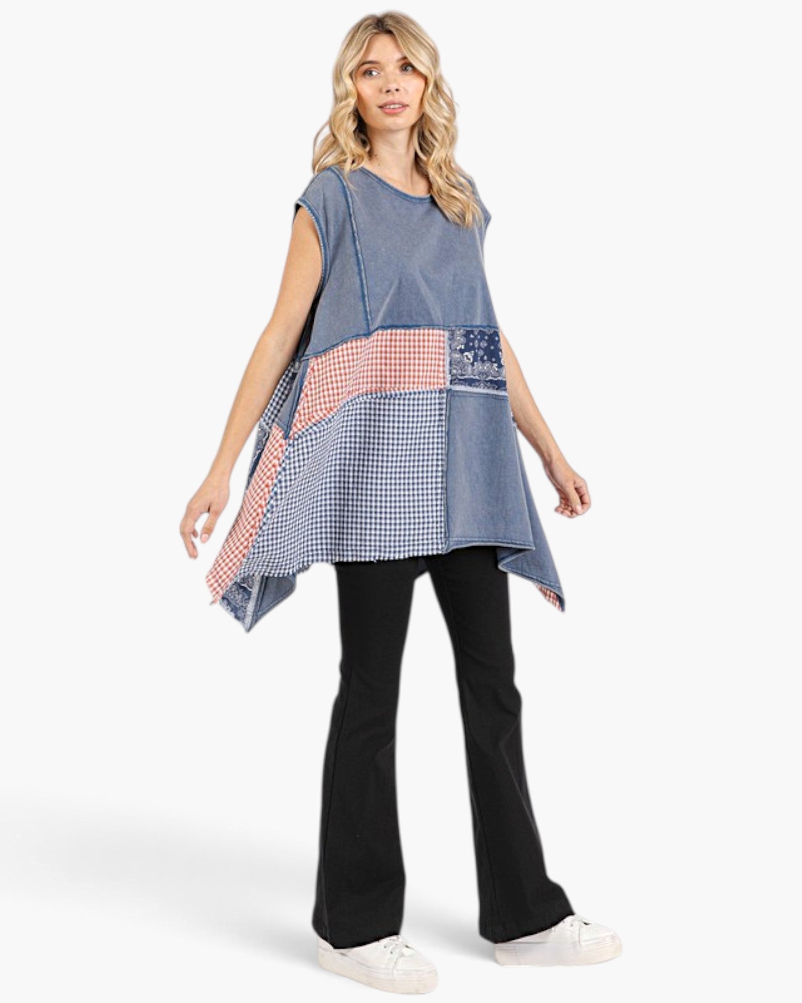 A woman modeling a blue patchwork tunic top with red and white gingham and checkered panels, paired with black pants and white sneakers, against a light gray studio background.
