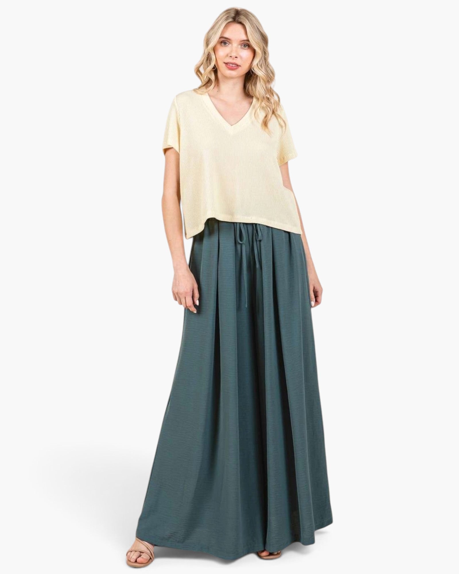 A woman modeling a cream short-sleeve V-neck top and dark teal wide-leg pleated pants against a plain white studio background.