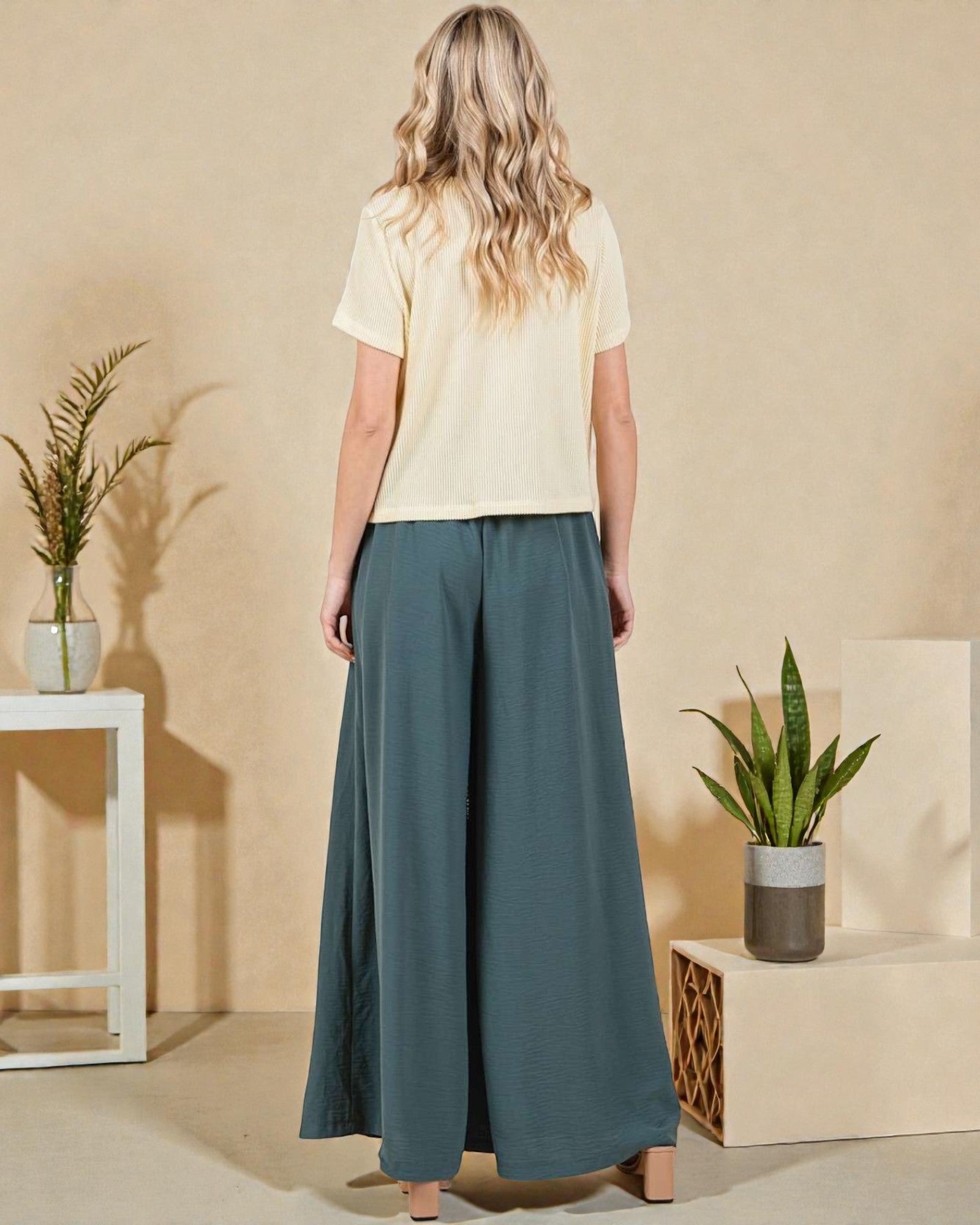 Woman wearing a light yellow top and teal pants standing in a minimalistic room with plants.