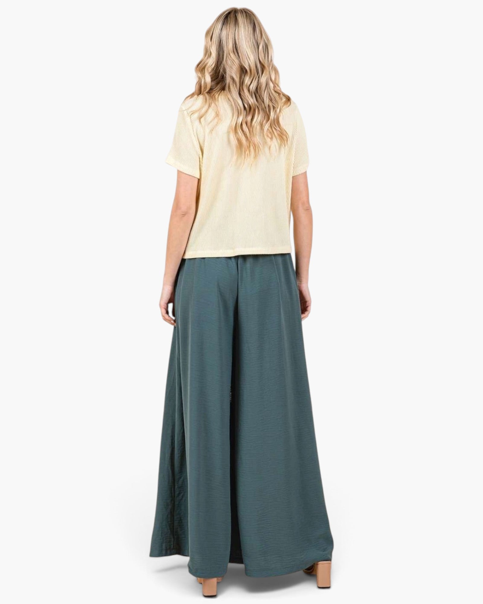 A woman shown from the back wearing a pale yellow short-sleeve top and dark teal wide-leg pants against a plain white studio background.