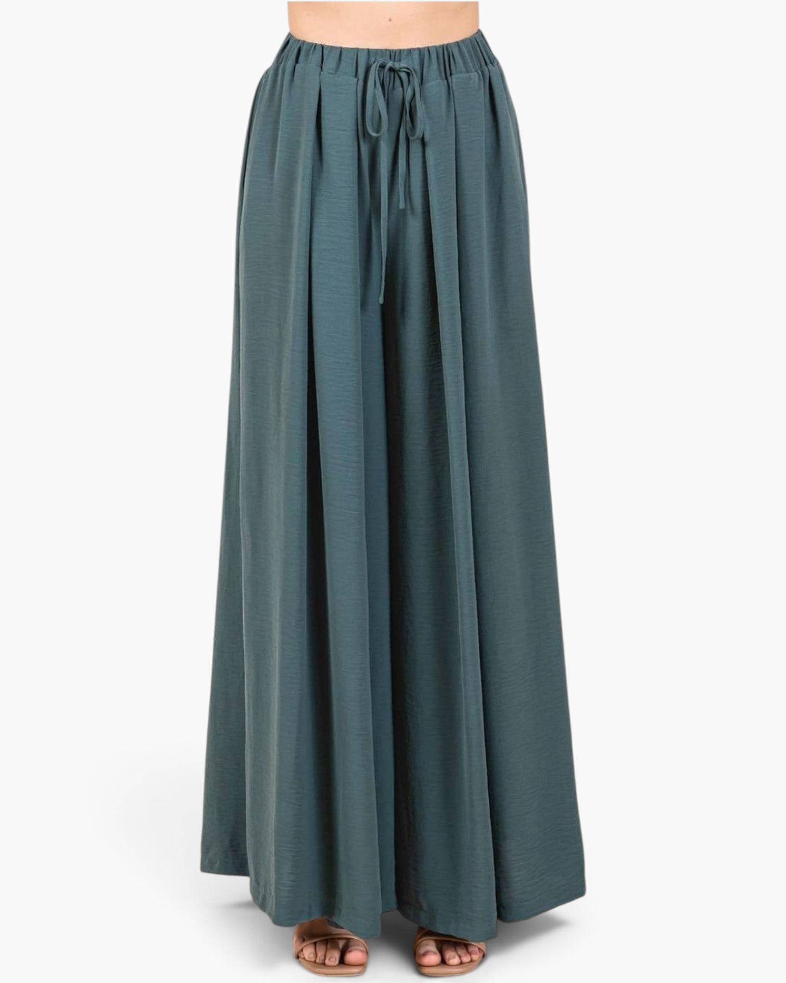 A person wearing a pair of wide-leg, floor-length pleated pants in a muted teal-green color, styled with brown strappy flat sandals against a plain light gray studio background.
