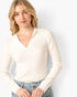 A woman wearing a cream ribbed long-sleeve V-neck top with a collar, paired with light-wash blue jeans, against a plain light gray background.