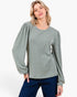A woman modeling a sage green long-sleeve top with dramatic balloon sleeves and dark blue jeans, photographed against a clean white studio background.