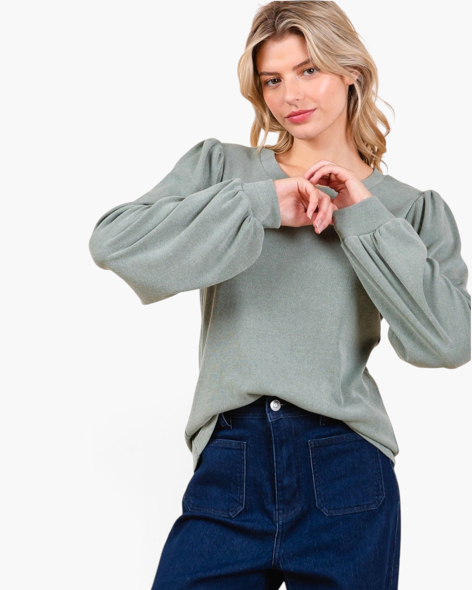 A woman wearing a sage green long-sleeve knit top with dramatic balloon sleeves and dark blue jeans, posing against a solid white studio background.