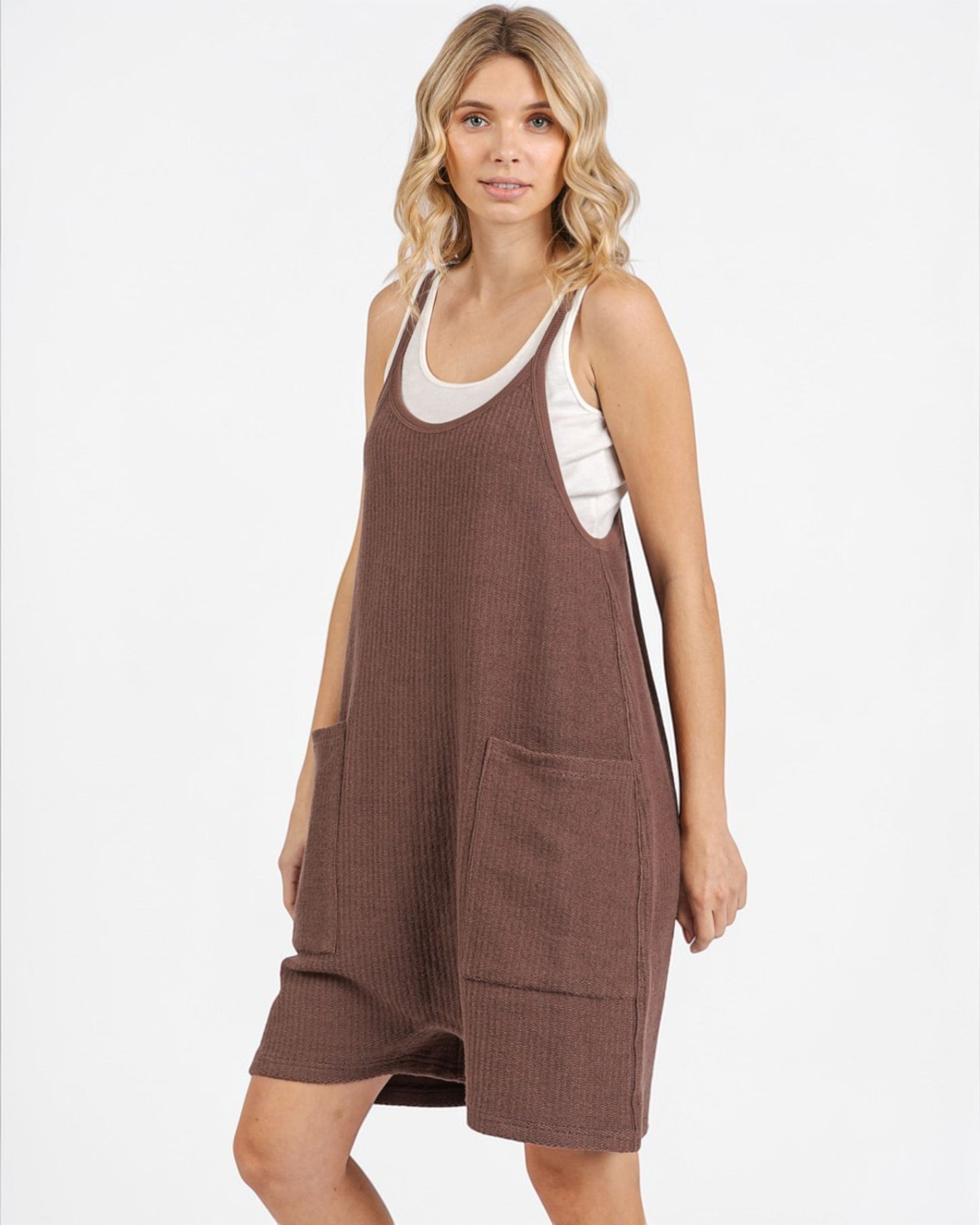 Woman wearing a brown overalls dress with a white top underneath on a white background