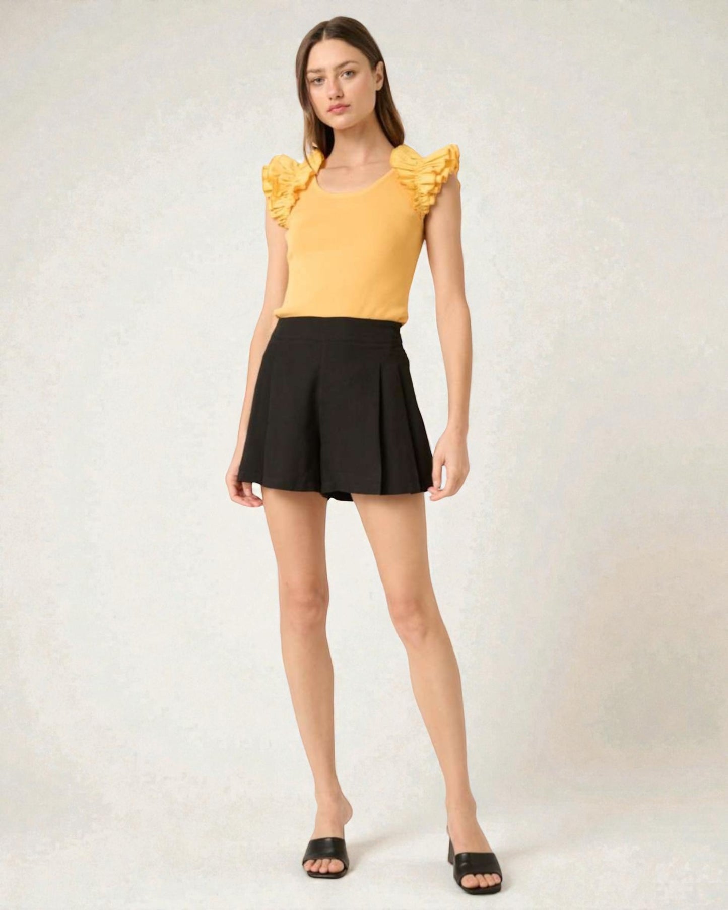 Woman wearing a yellow top with ruffled sleeves and black shorts on a white background
