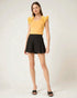 Woman wearing a yellow top with ruffled sleeves and black shorts on a white background