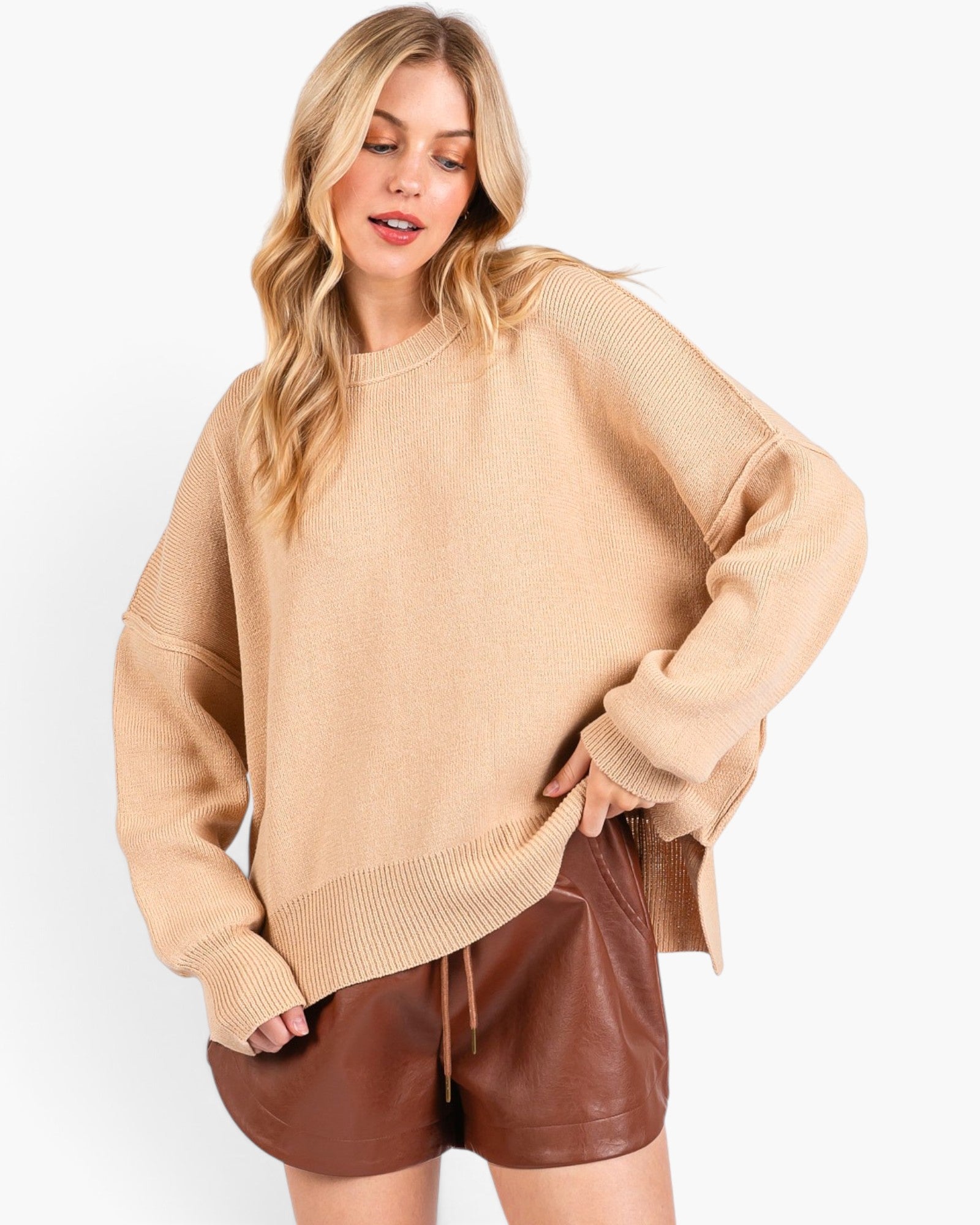 A woman modeling an oversized beige knit crewneck sweater paired with brown faux-leather shorts against a plain white studio background.