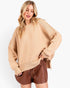 A woman modeling an oversized beige knit crewneck sweater paired with brown faux-leather shorts against a plain white studio background.