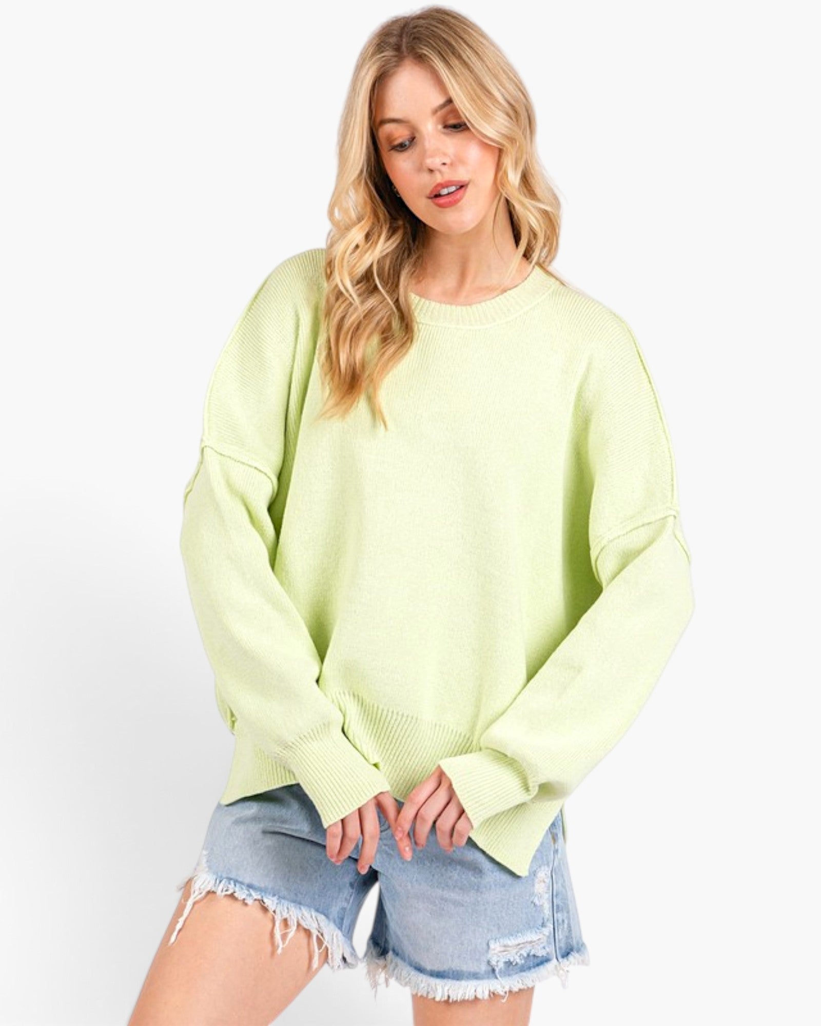 A woman wearing an oversized light lime green knit sweater and light-wash frayed denim shorts, standing against a plain white studio background.