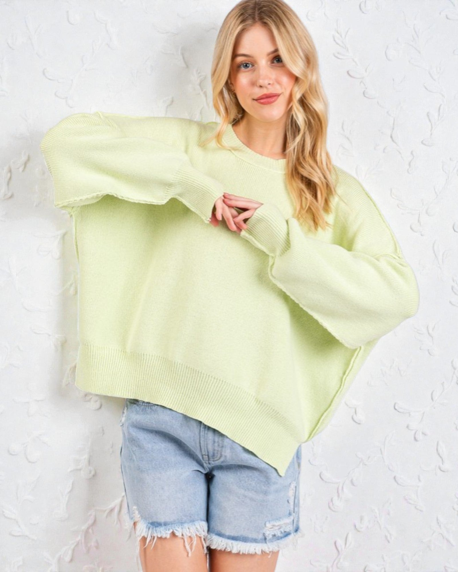 Woman wearing a light green sweater and denim shorts against a white textured wall.