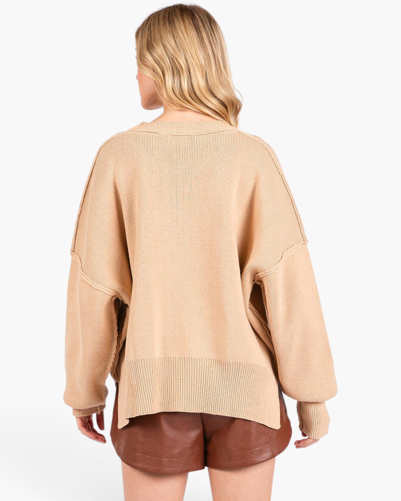 A woman shown from the back wearing a loose beige knit sweater and brown shorts against a plain light gray studio background.