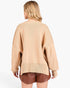A woman shown from the back wearing a loose beige knit sweater and brown shorts against a plain light gray studio background.