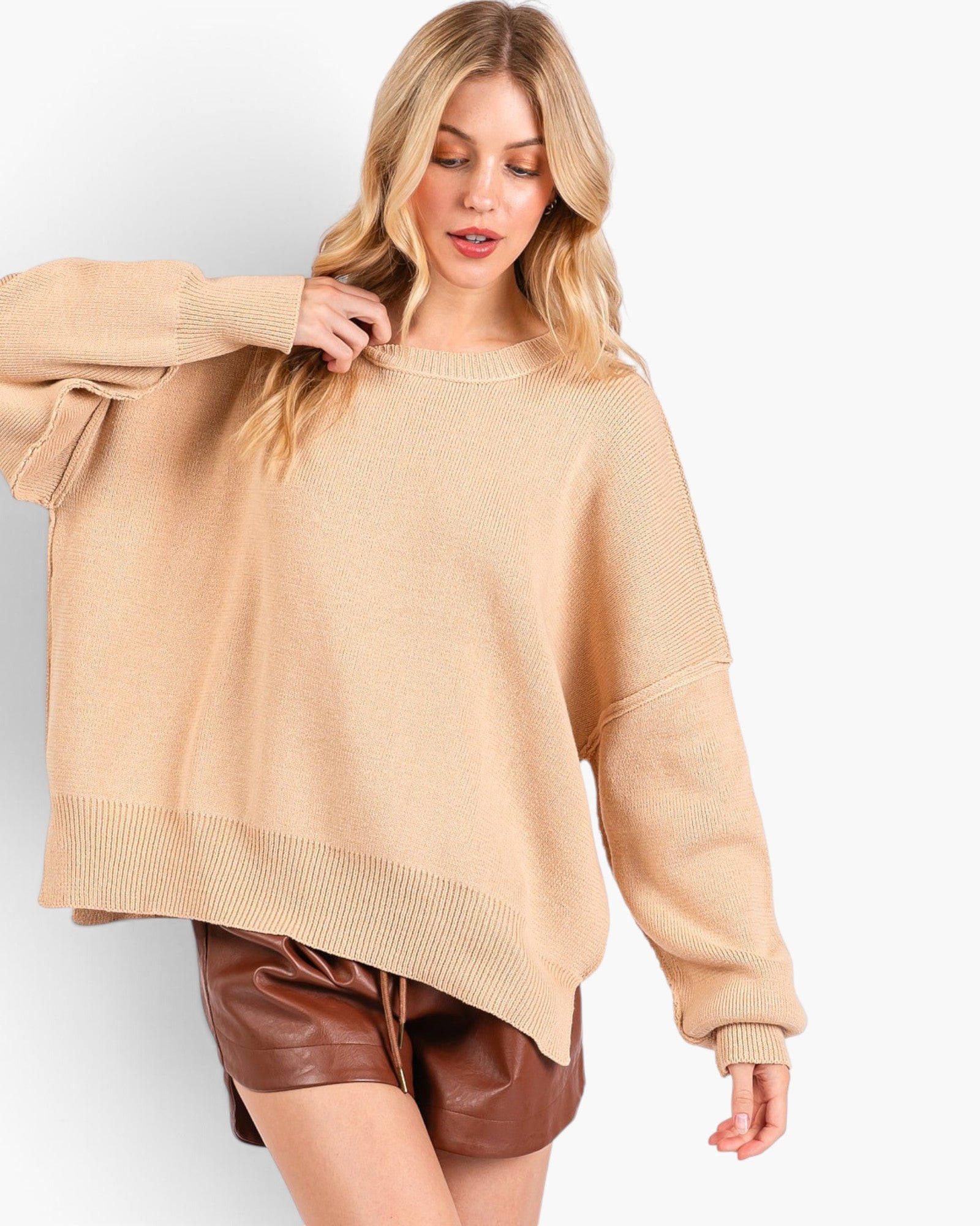 A woman wearing a loose, oversized beige knit sweater and brown faux-leather shorts against a clean white studio background.