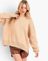 A woman wearing a loose, oversized beige knit sweater and brown faux-leather shorts against a clean white studio background.