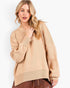 A woman wearing a beige crewneck knit sweater with long sleeves and ribbed cuffs, hem, and collar, paired with brown leather pants, against a plain white studio background.