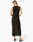 A woman viewed from the back wearing a sleeveless black midi dress with a subtle textured pattern and a sheer mesh panel near the lower back, paired with black mule heels against a light gray studio background.