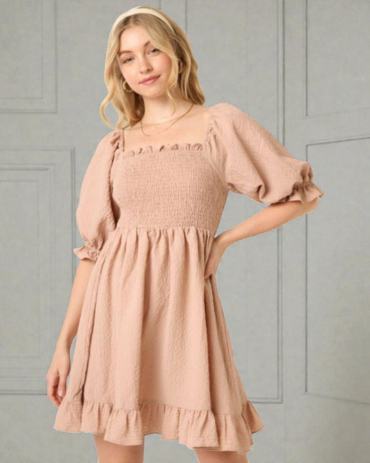 Woman wearing a beige dress with ruffled sleeves and hem against a gray paneled wall.