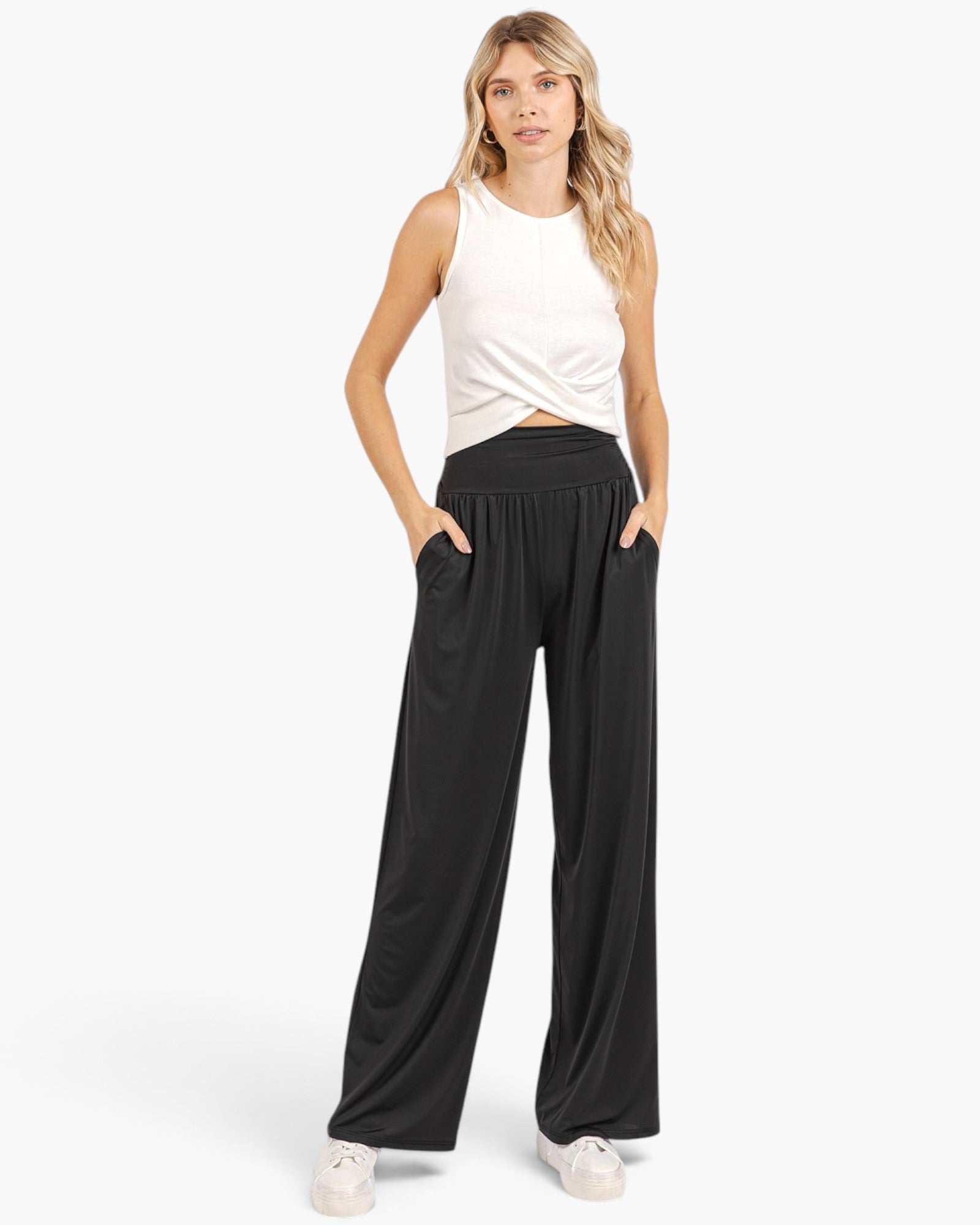 A woman modeling black wide-leg pants with a white sleeveless top and white sneakers, standing against a light gray studio background.