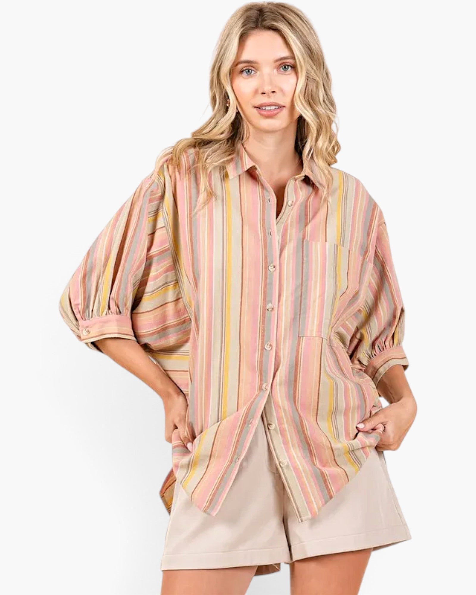 A woman modeling a loose, short-sleeve button-up shirt with vertical stripes in pink, yellow, green, and beige, paired with beige shorts against a plain white background.