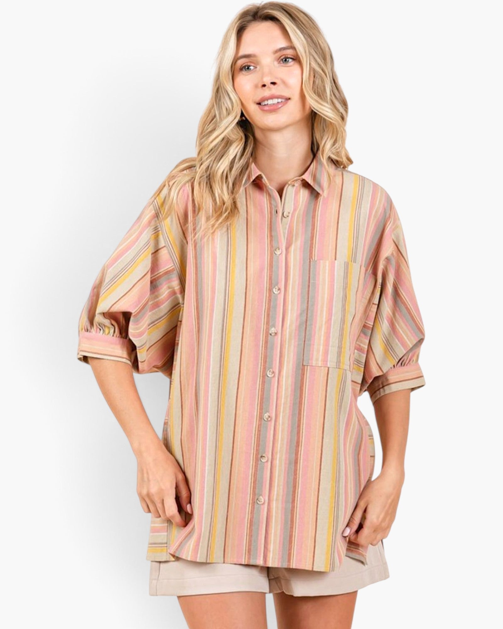 A woman modeling a loose, oversized button-up shirt with vertical stripes in pink, yellow, beige, and muted green, paired with beige shorts against a plain light gray studio background.