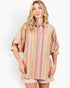 A woman modeling a loose, oversized button-up shirt with vertical stripes in pink, yellow, beige, and muted green, paired with beige shorts against a plain light gray studio background.