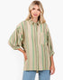 A woman modeling a loose-fitting button-up shirt with vertical stripes in sage green, beige, and cream, paired with light blue jeans against a plain white background.