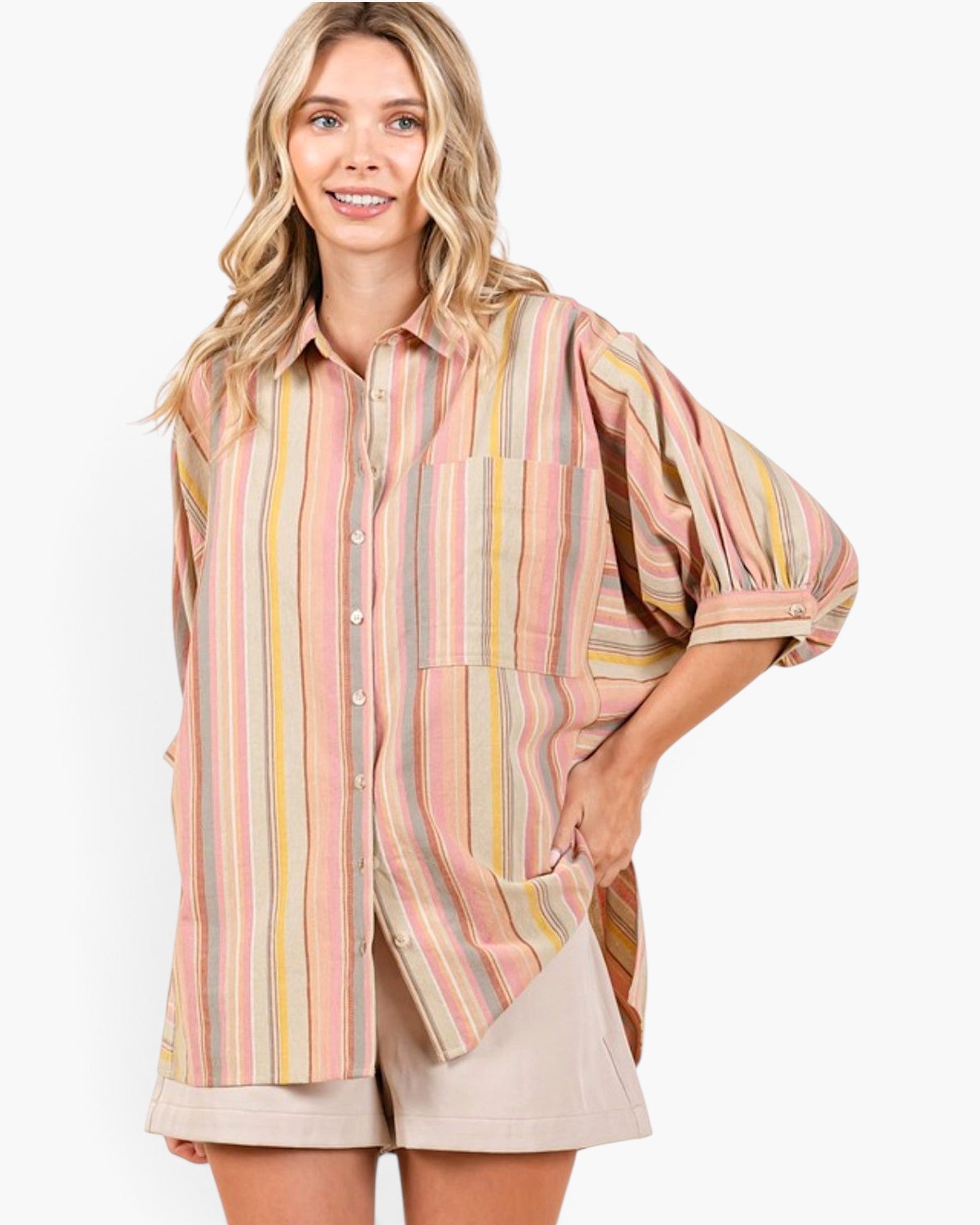 A woman modeling a loose, oversized button-up shirt with vertical pastel stripes in pink, yellow, green, and beige, paired with light beige shorts against a plain white background.