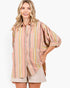 A woman modeling a loose, oversized button-up shirt with vertical pastel stripes in pink, yellow, green, and beige, paired with light beige shorts against a plain white background.