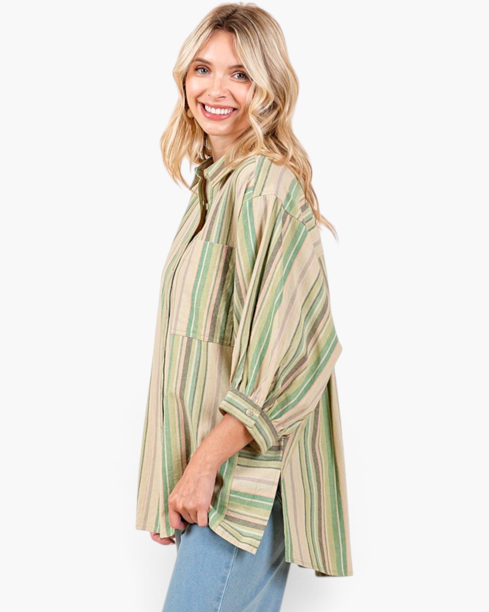 A woman wearing a loose, oversized striped button-up shirt in beige, green, and brown tones, paired with light blue jeans, photographed against a clean white studio background.