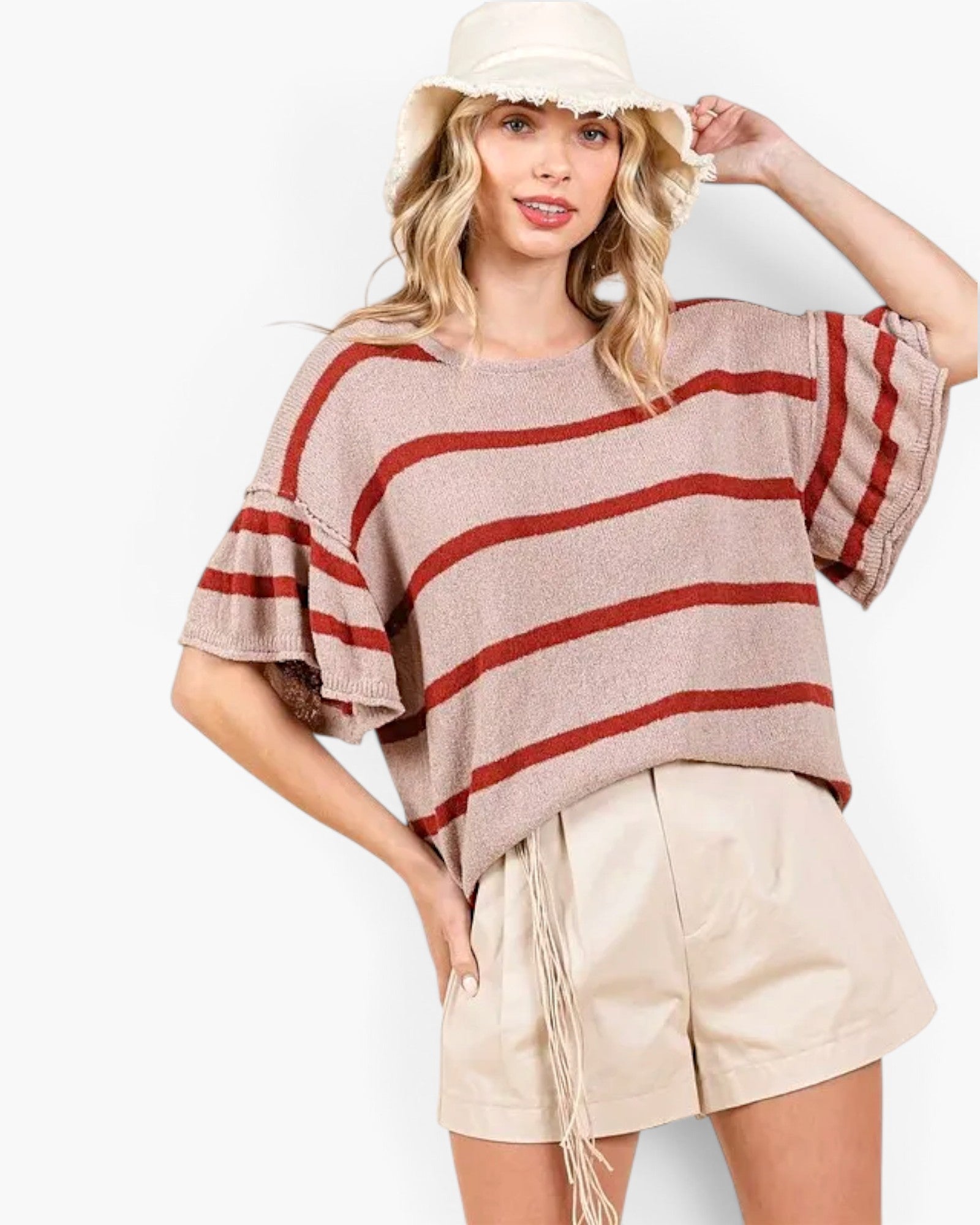 A woman modeling a loose knit top with horizontal rust-red stripes on a light taupe base, paired with beige shorts and a cream bucket hat against a plain white background.