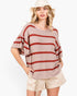 A woman wearing a beige knit top with horizontal rust-red stripes, paired with beige shorts and a cream bucket hat, against a plain white background.