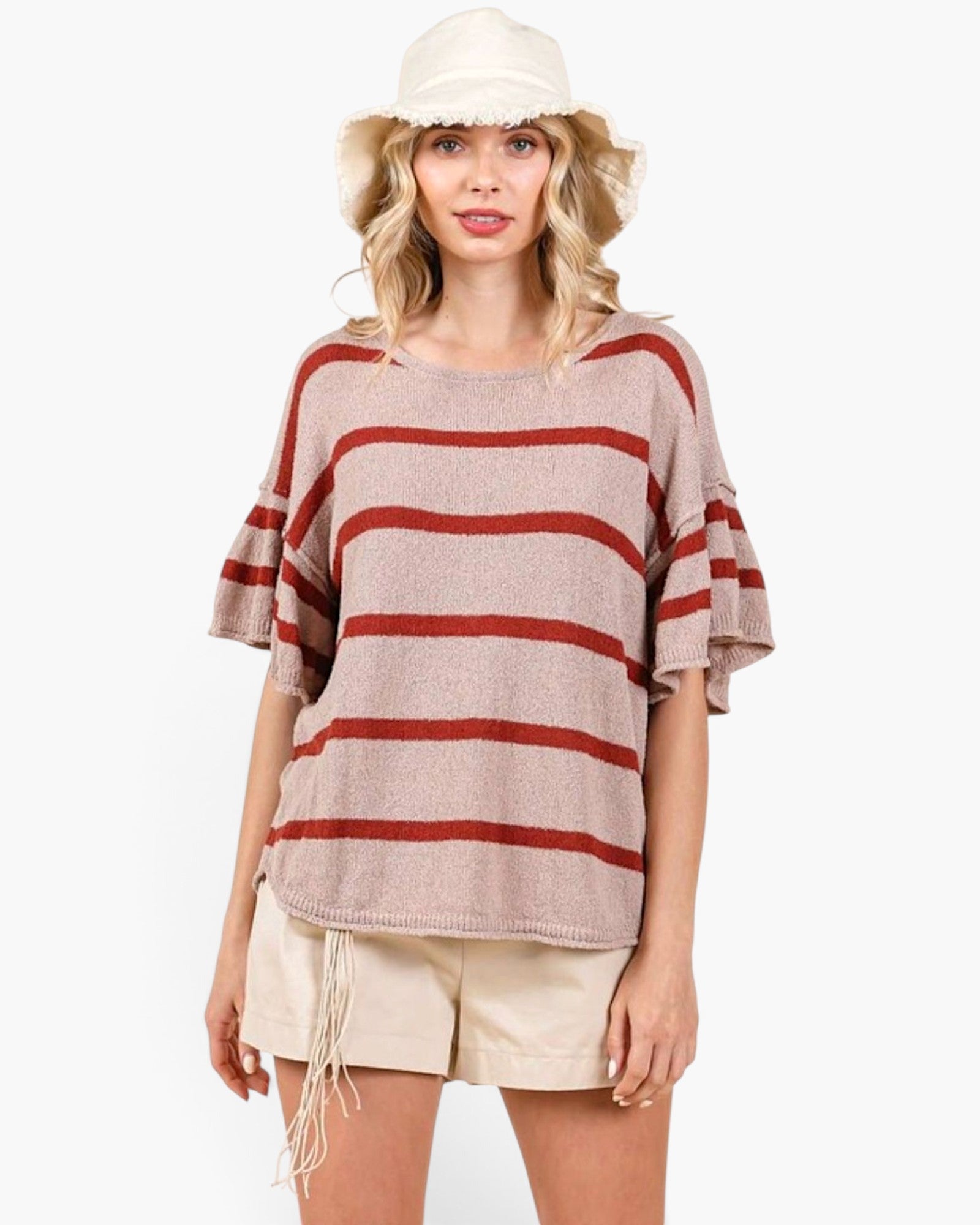 A woman modeling a loose, short-sleeve knit top with horizontal rust-red stripes on a light beige background, paired with cream shorts and a cream bucket hat against a plain white studio background.
