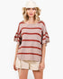 A woman modeling a loose, short-sleeve knit top with horizontal rust-red stripes on a light beige background, paired with cream shorts and a cream bucket hat against a plain white studio background.