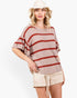 A woman modeling a loose knit top with horizontal rust-red stripes on a light beige base, paired with beige shorts and a cream hat against a plain white background.