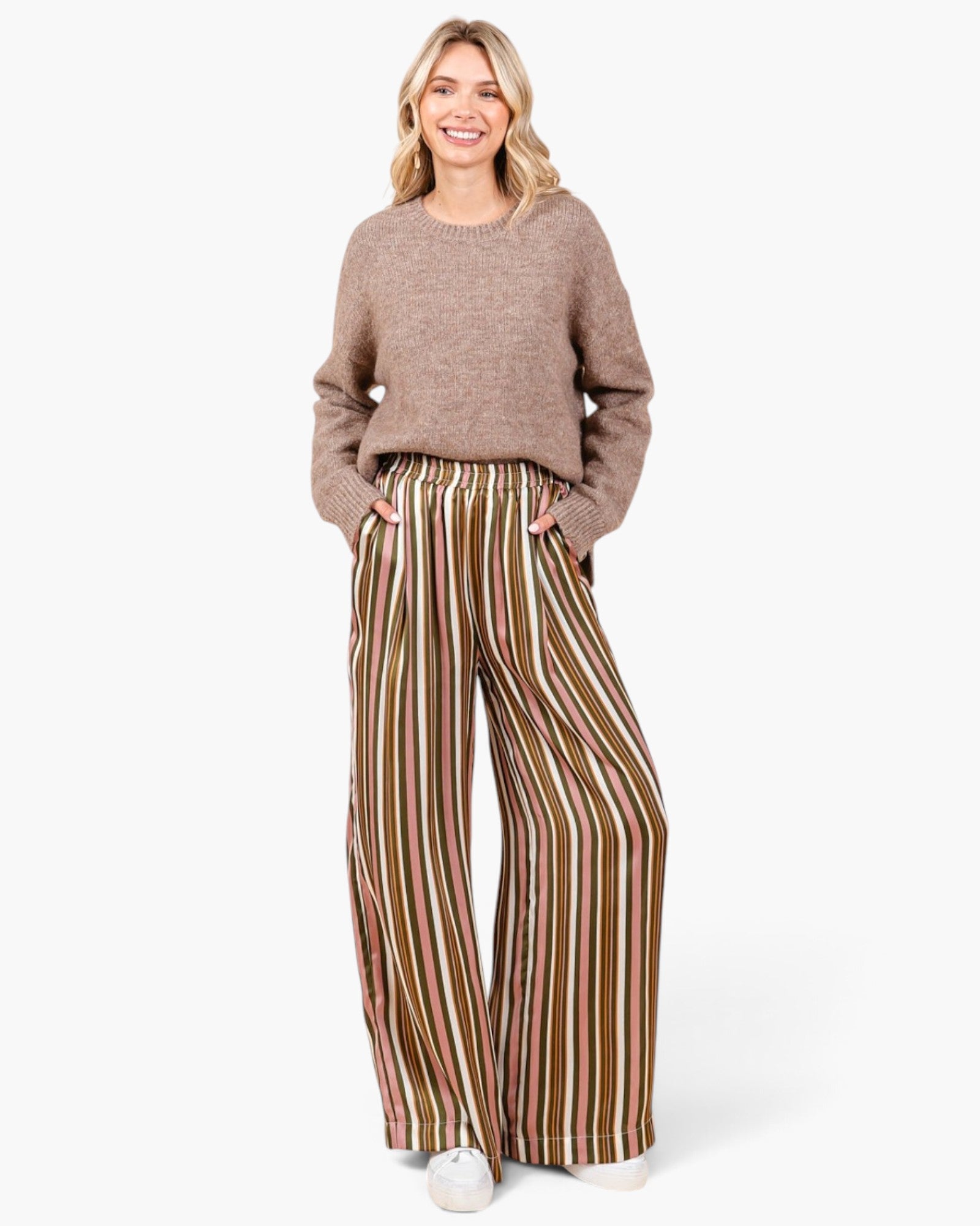 A woman modeling a cozy taupe knit sweater and wide-leg striped trousers with vertical stripes in brown, gold, and cream, standing against a plain white studio background.