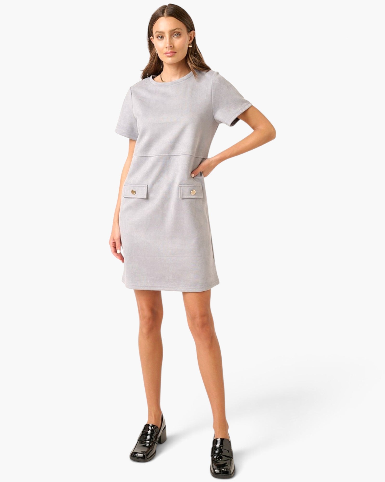 A woman modeling a light gray short-sleeve dress with two front flap pockets featuring gold-tone buttons, paired with black loafers, against a plain white studio background.