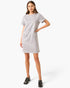 A woman modeling a light gray short-sleeve dress with two front flap pockets featuring gold-tone buttons, paired with black loafers, against a plain white studio background.