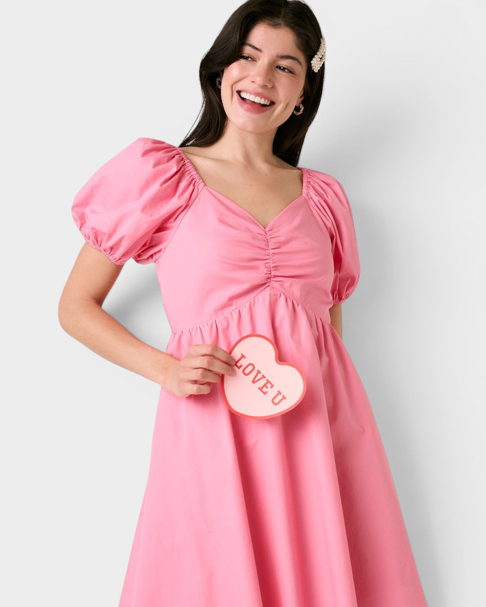 Woman wearing a pink dress holding a heart-shaped 'LOVE' sign against a white background