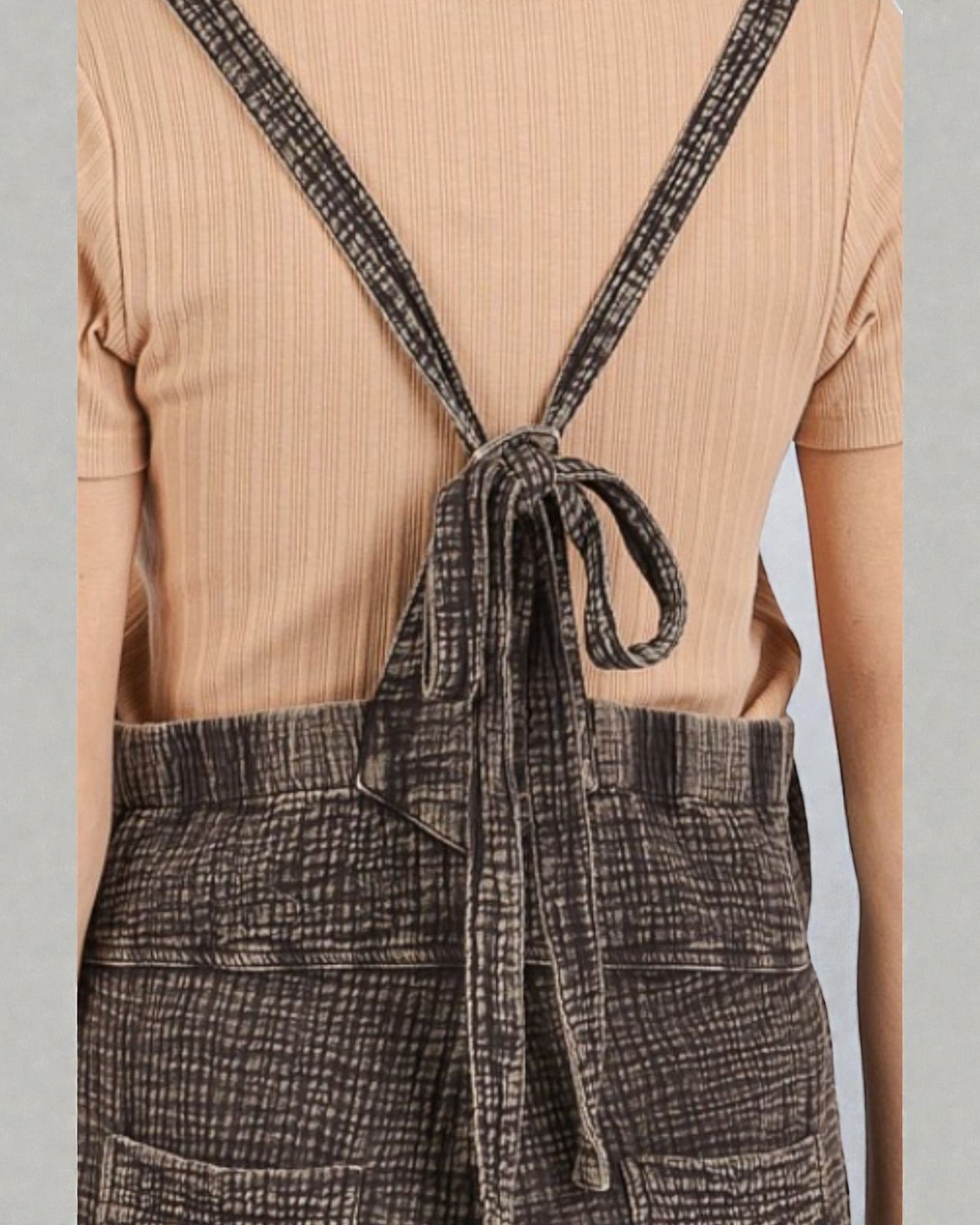 Person wearing a beige ribbed top and plaid overalls with a gray background