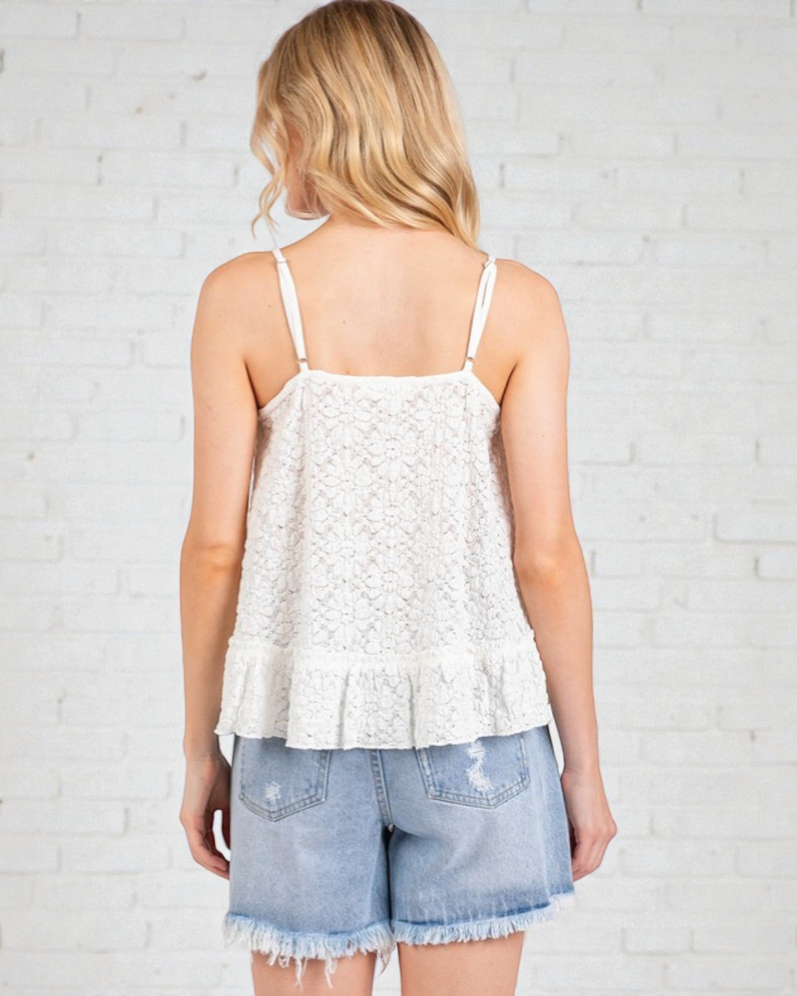 Woman wearing a white lace top and denim shorts against a light brick wall.