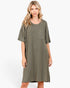 A woman modeling an oversized short-sleeve dress with a vertical textured stripe pattern in olive green and beige, featuring a single chest pocket, set against a plain white studio background.