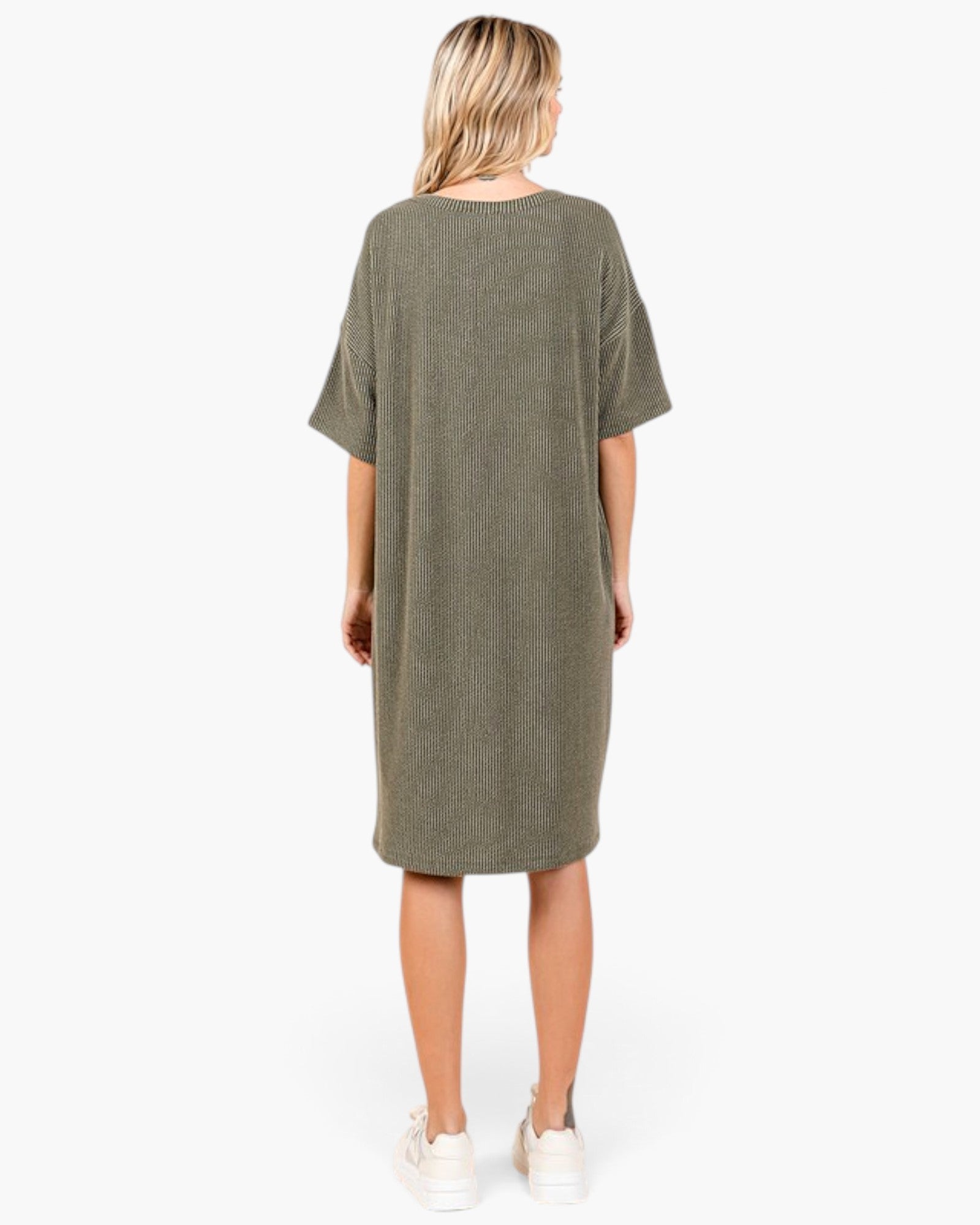 A woman viewed from the back wearing an oversized short-sleeve knit dress in olive green, paired with white sneakers, against a plain white studio background.