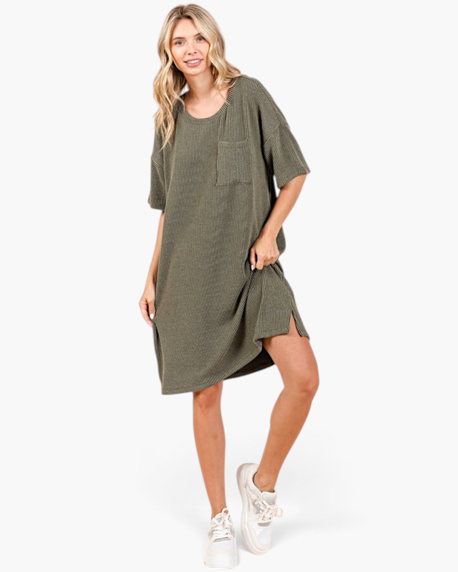 A woman modeling an oversized short-sleeve olive green tunic dress with a chest pocket, paired with white sneakers, against a plain white studio background.